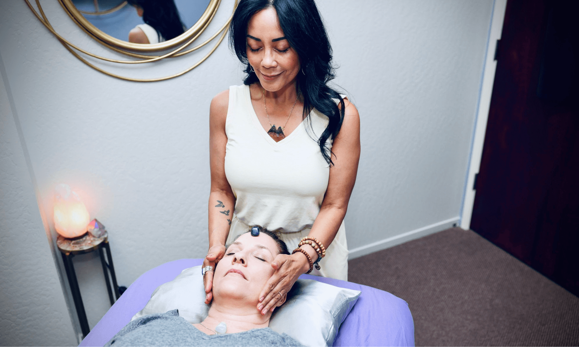 A beautiful woman with long dark hair and tan skin, Deanna Jaromay, practicing Reiki, energy work, with her hands on the temples of another woman laying on a table.