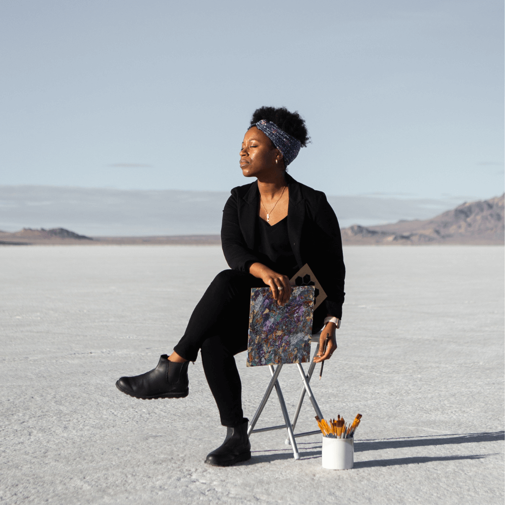Andrea Hardeman sitting on a chair out in the white Salt Flats of Utah, mountains in the background, holding her artwork and looking off into the distance.
