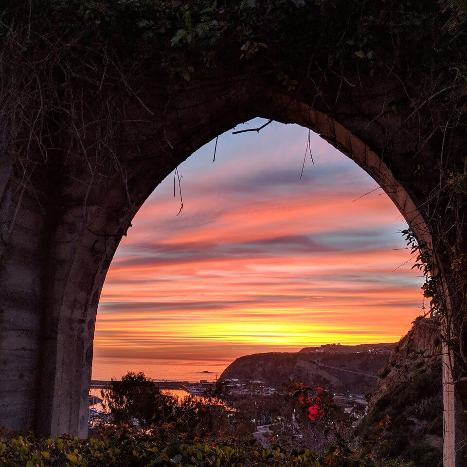 Stunning sunset view through an archway at Doheny Beach in Dana Point, CA, featuring vibrant orange and pink hues over the Pacific Ocean, with coastal cliffs and marina visible in the distance.